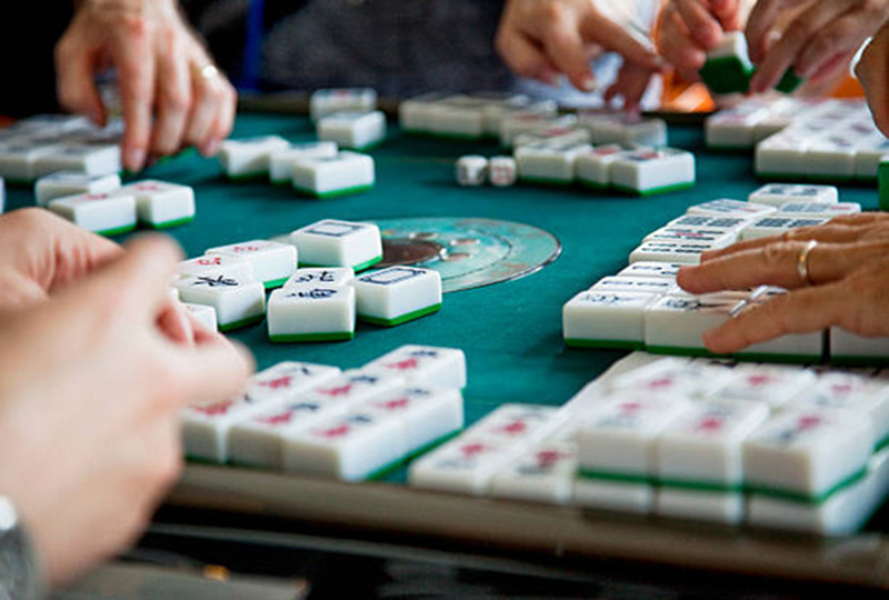 Four pair of hands playing with white and green tiles at a table with green felt cover