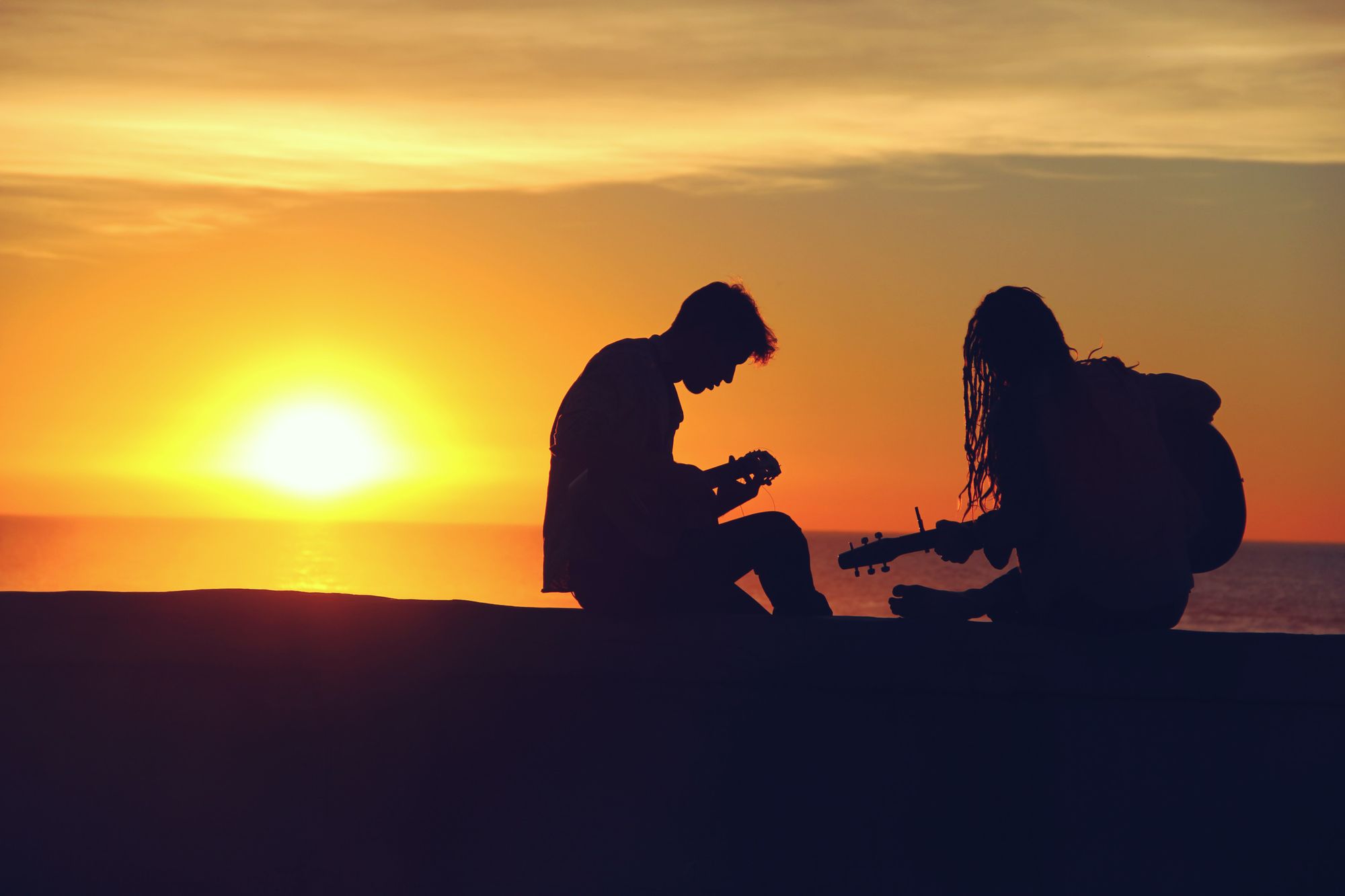 Two people playing the guitar on a beach while the sun sets